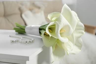 Beautiful calla lily flowers tied with ribbon and jewelry on white chest of drawers indoors, closeup