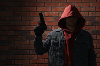Thief in hoodie with gun against red brick wall. Space for text