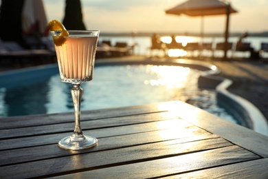 Photo of Glass of fresh summer cocktail on wooden table near swimming pool outdoors at sunset. Space for text