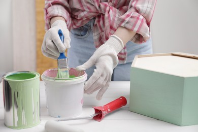 Photo of Woman dipping brush into bucket of green paint at white table indoors, closeup