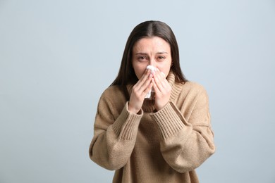 Young woman with tissue suffering from runny nose on light grey background