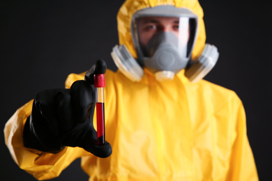 Man in chemical protective suit holding test tube of blood sample against black background, focus on hand. Virus research