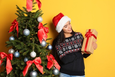 Beautiful young woman in Santa hat with gift box near Christmas tree on color background