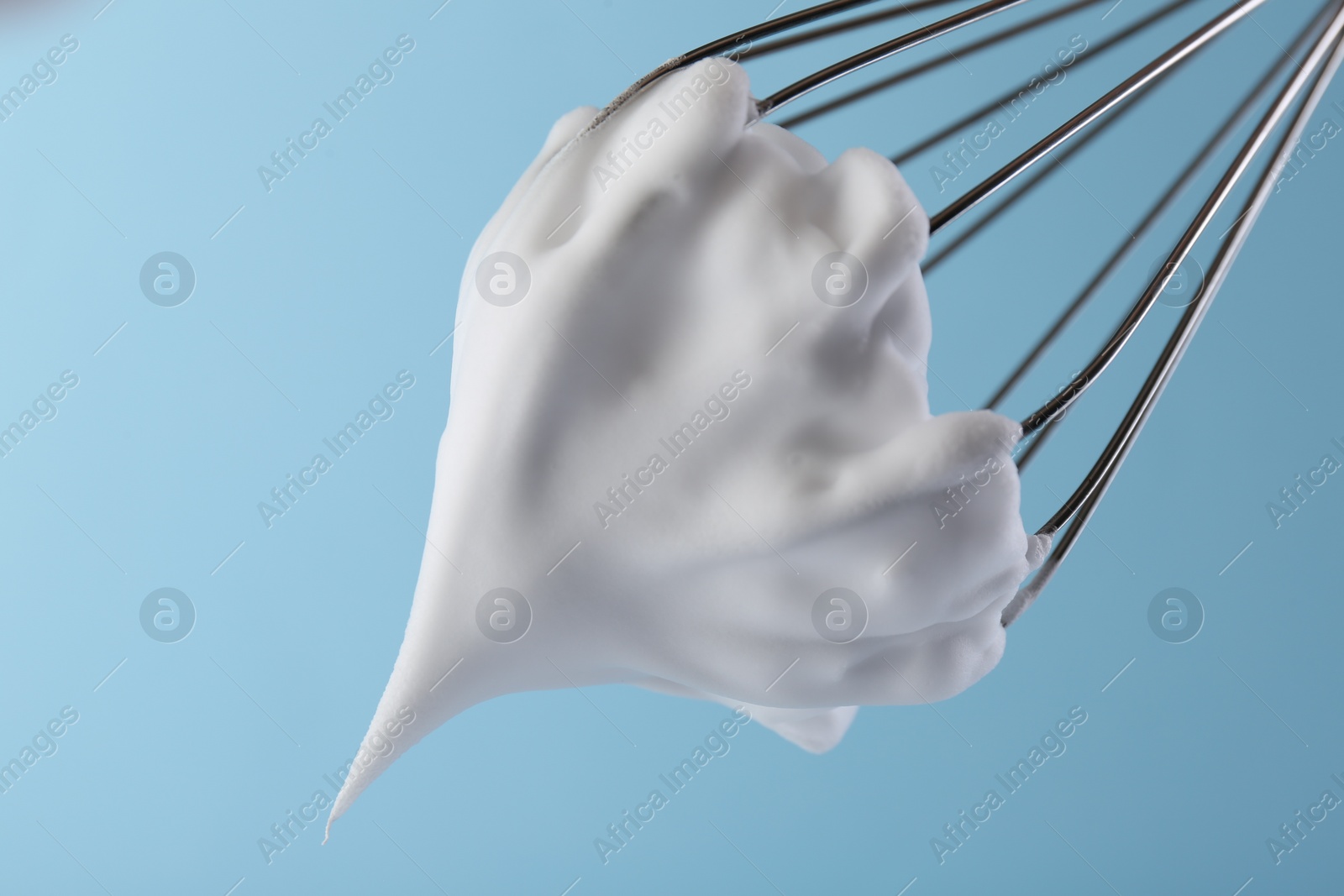 Photo of Whisk with whipped cream on light blue background, closeup