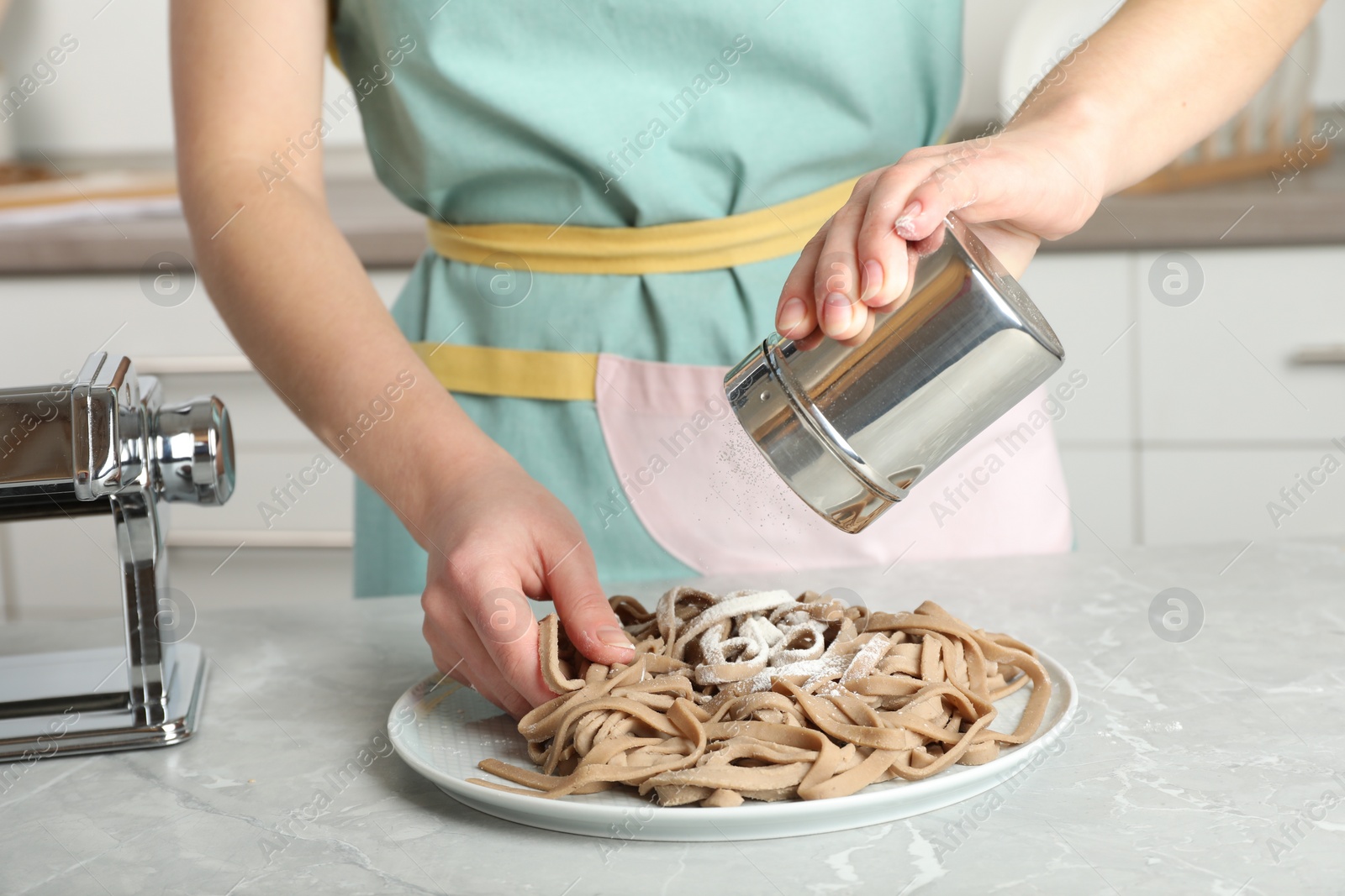 Photo of Woman sprinkling raw soba (buckwheat noodles) with flour at grey table in kitchen, closeup