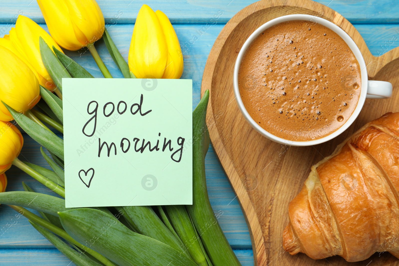 Photo of Cup of aromatic coffee with croissant, beautiful yellow tulips and Good Morning note on light blue wooden table, flat lay