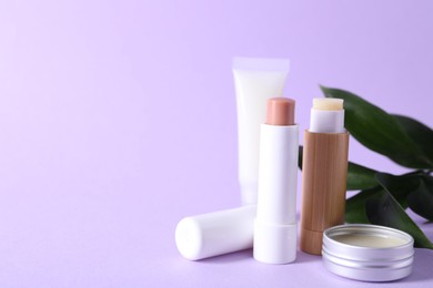 Photo of Different lip balms and leaves on lilac background, closeup. Space for text