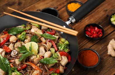 Photo of Stir-fry. Tasty noodles with meat in wok, chopsticks and ingredients on wooden table, closeup