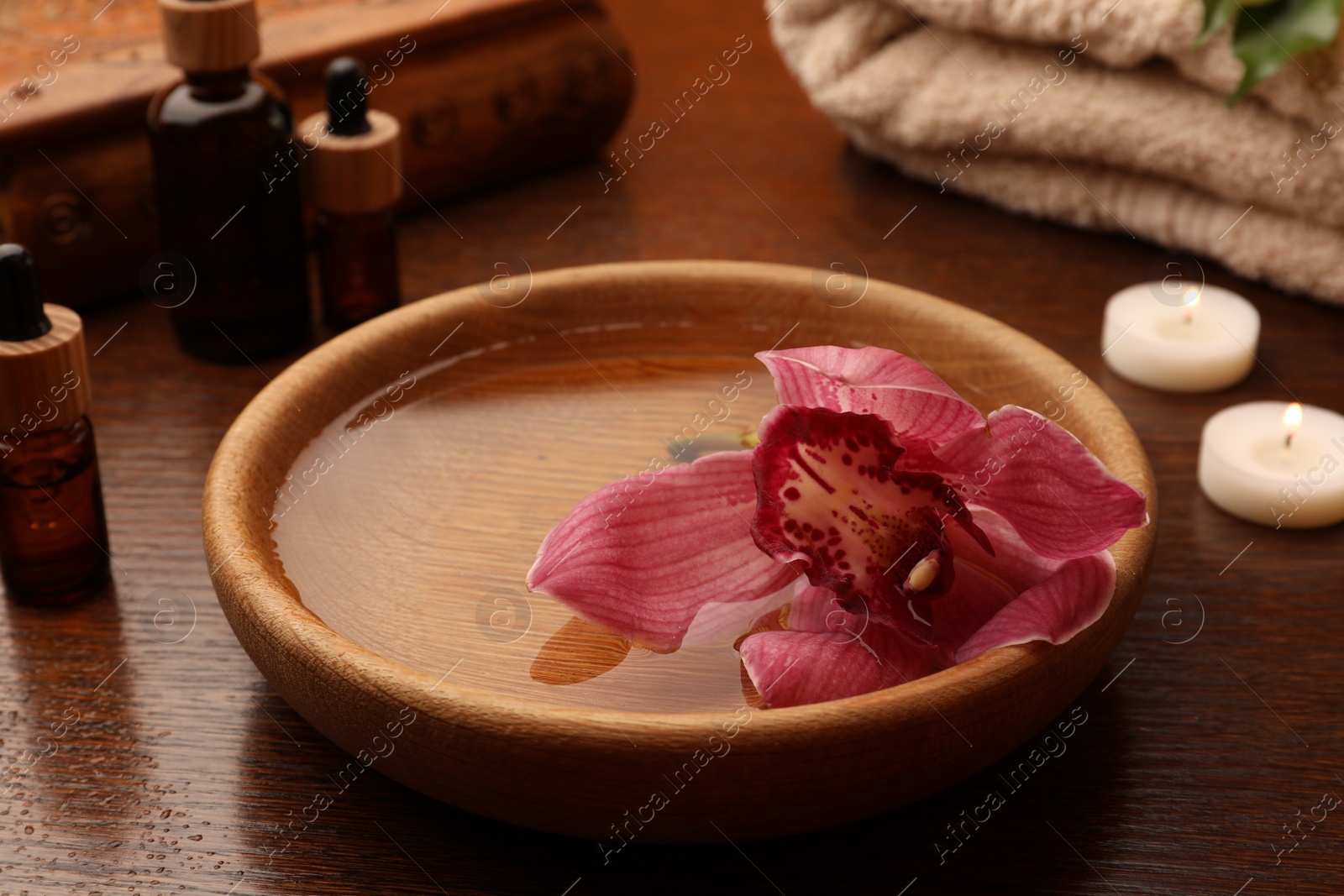 Photo of Water with beautiful flower in bowl, bottles of essential oil and candles on wooden table. Aromatherapy treatment