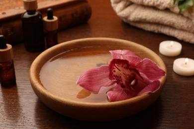 Water with beautiful flower in bowl, bottles of essential oil and candles on wooden table. Aromatherapy treatment