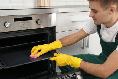 Male janitor cleaning oven tray with sponge in kitchen