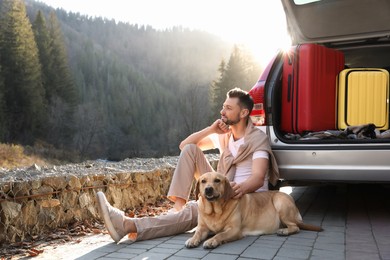 Photo of Man and adorable dog sitting near car in mountains. Traveling with pet