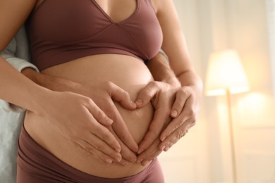 Photo of Pregnant young woman making heart with hands on belly and husband near her at home, closeup