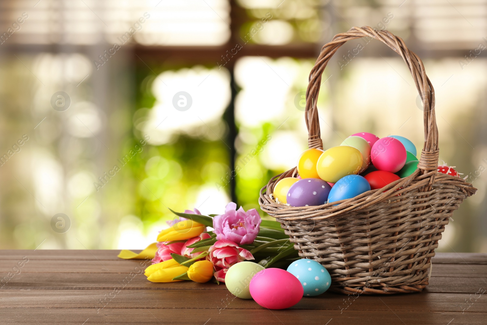 Image of Colorful Easter eggs in wicker basket and tulips on wooden table indoors, space for text
