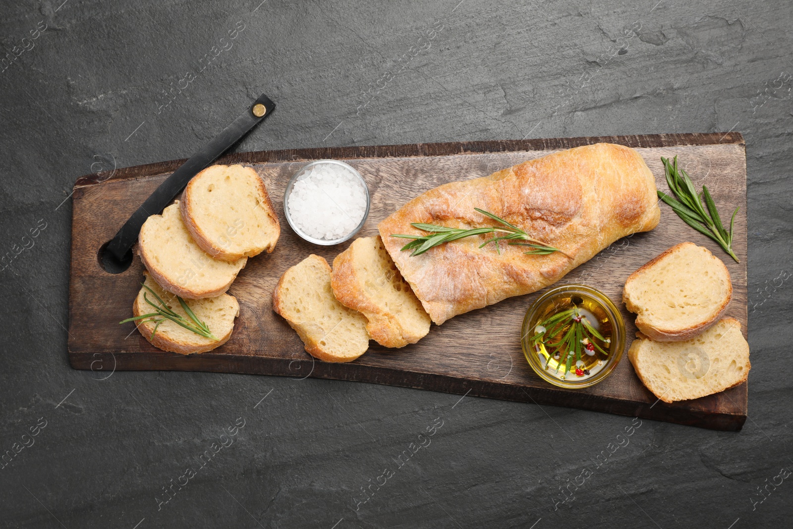Photo of Delicious French baguette with rosemary, oil and salt on black table, top view
