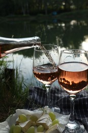 Photo of Pouring delicious rose wine into glass on picnic blanket near lake, closeup