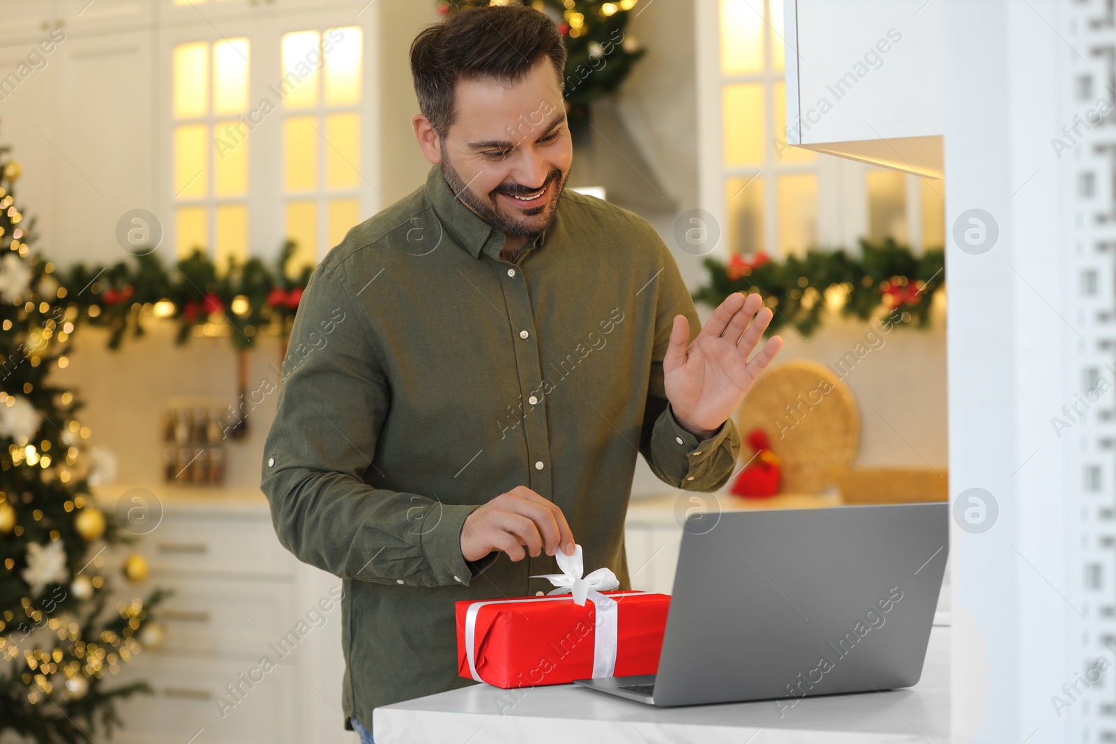 Photo of Celebrating Christmas online with exchanged by mail presents. Happy man opening gift box during video call on laptop in kitchen