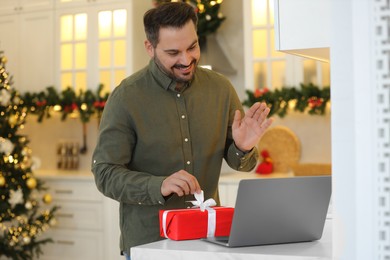 Celebrating Christmas online with exchanged by mail presents. Happy man opening gift box during video call on laptop in kitchen