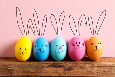 Colorful eggs as Easter bunnies on wooden table against pink background