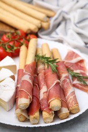 Plate of delicious grissini sticks with prosciutto, cheese and tomatoes on light grey table, closeup
