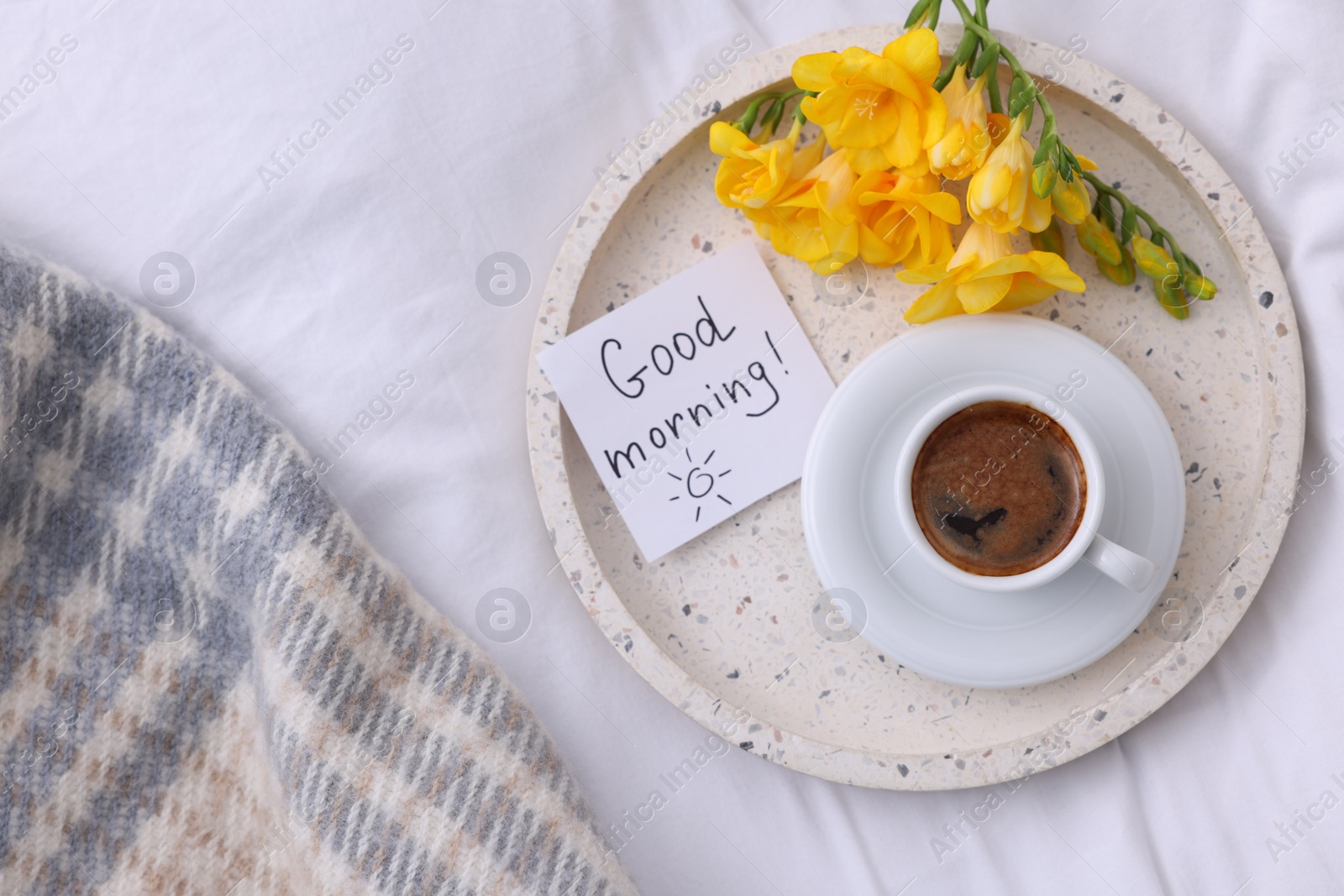 Photo of Cup of coffee, flowers and card with phrase GOOD MORNING! on bed, flat lay