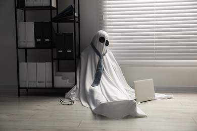 Photo of Overworked ghost. Man in white sheet with laptop on floor in office