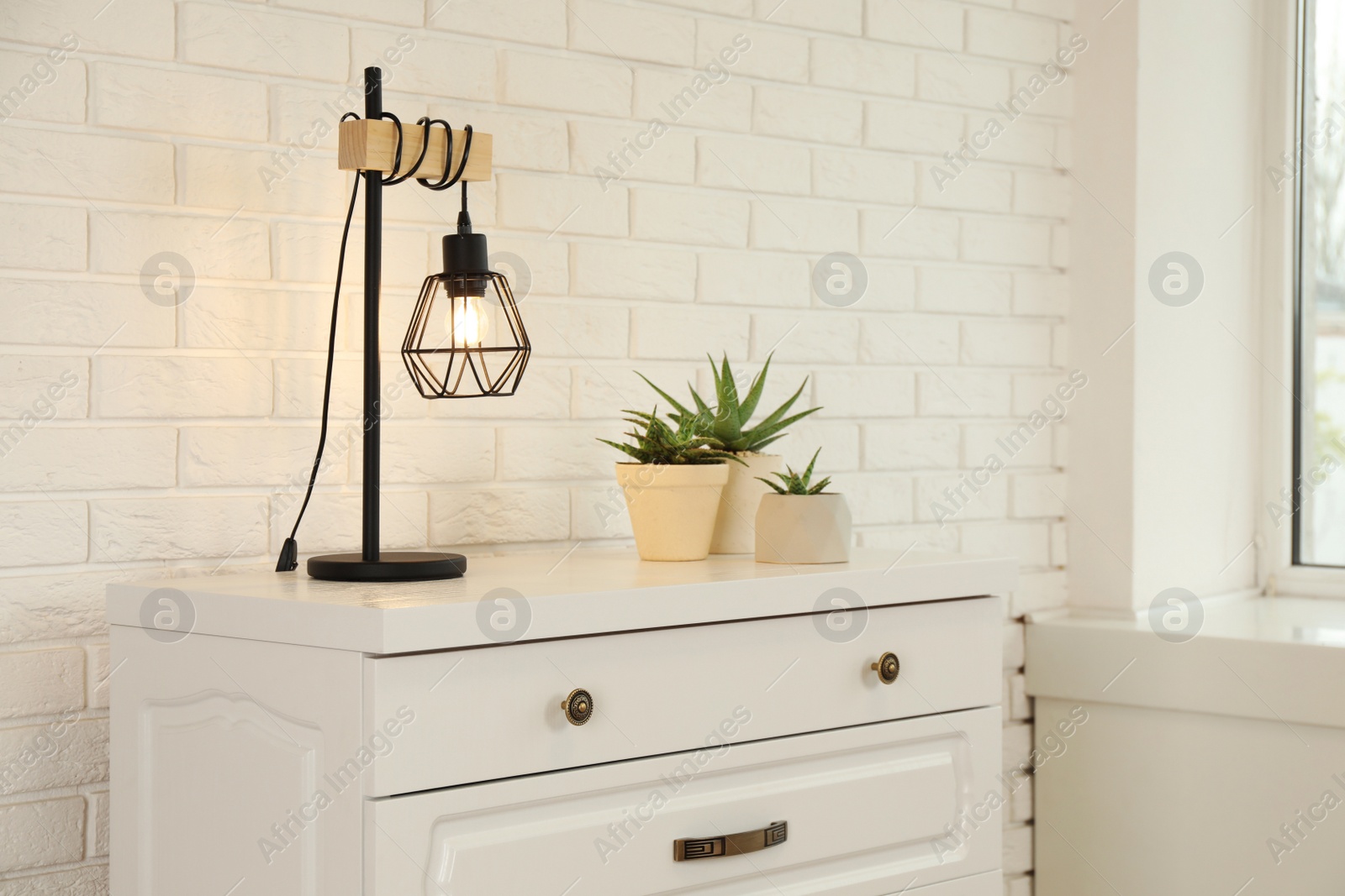 Photo of Modern chest of drawers with houseplants and lamp near white brick wall