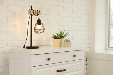 Photo of Modern chest of drawers with houseplants and lamp near white brick wall