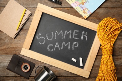 Chalkboard with text SUMMER CAMP and camping equipment on wooden background, flat lay