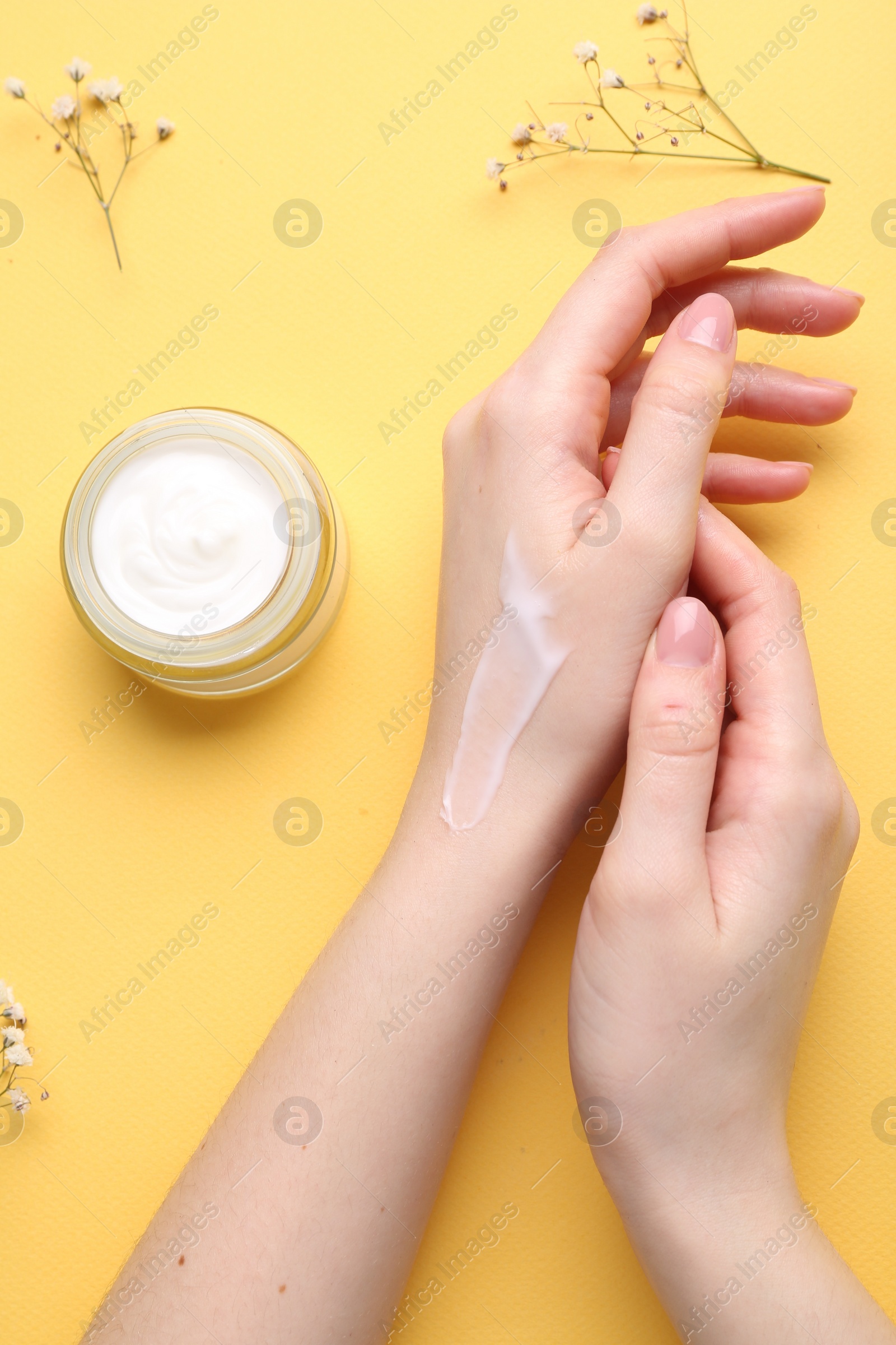 Photo of Woman applying hand cream and flowers on yellow background, top view