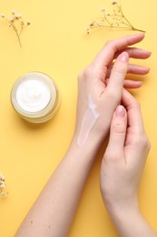 Photo of Woman applying hand cream and flowers on yellow background, top view