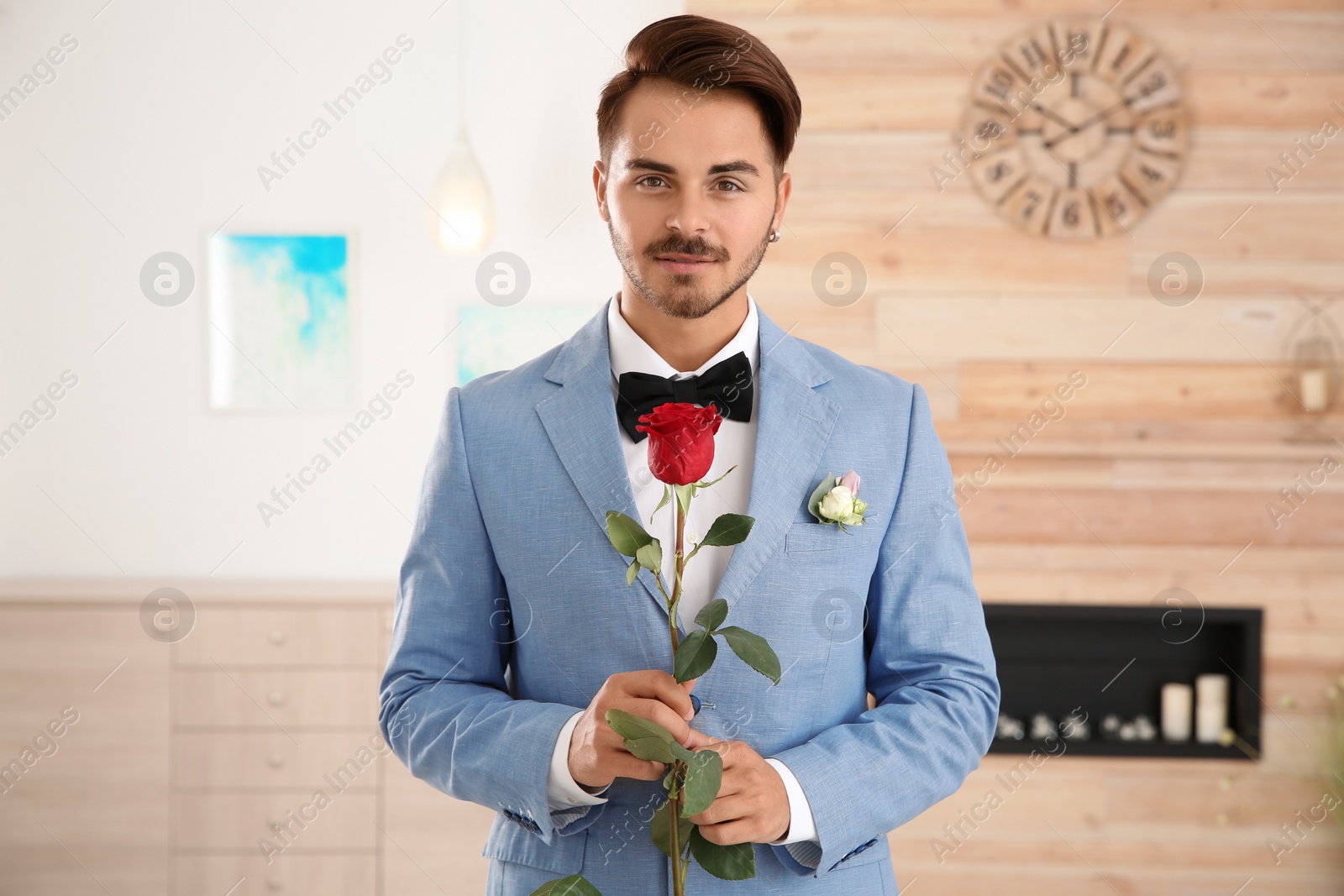 Photo of Handsome man in formal wear holding red rose indoors