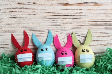 Photo of Dyed eggs with bunny ears in protective masks on wooden background, flat lay and space for text. Easter holiday during COVID-19 quarantine