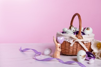 Wicker basket with festively decorated Easter eggs, bunny and beautiful tulips on white marble table against pink background. Space for text