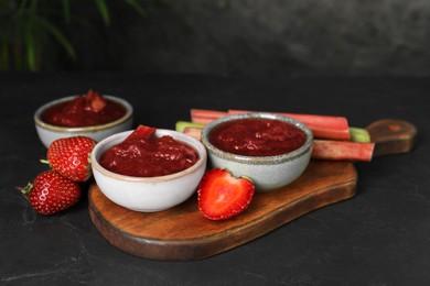 Photo of Tasty rhubarb jam in bowls, stems and strawberries on dark textured table