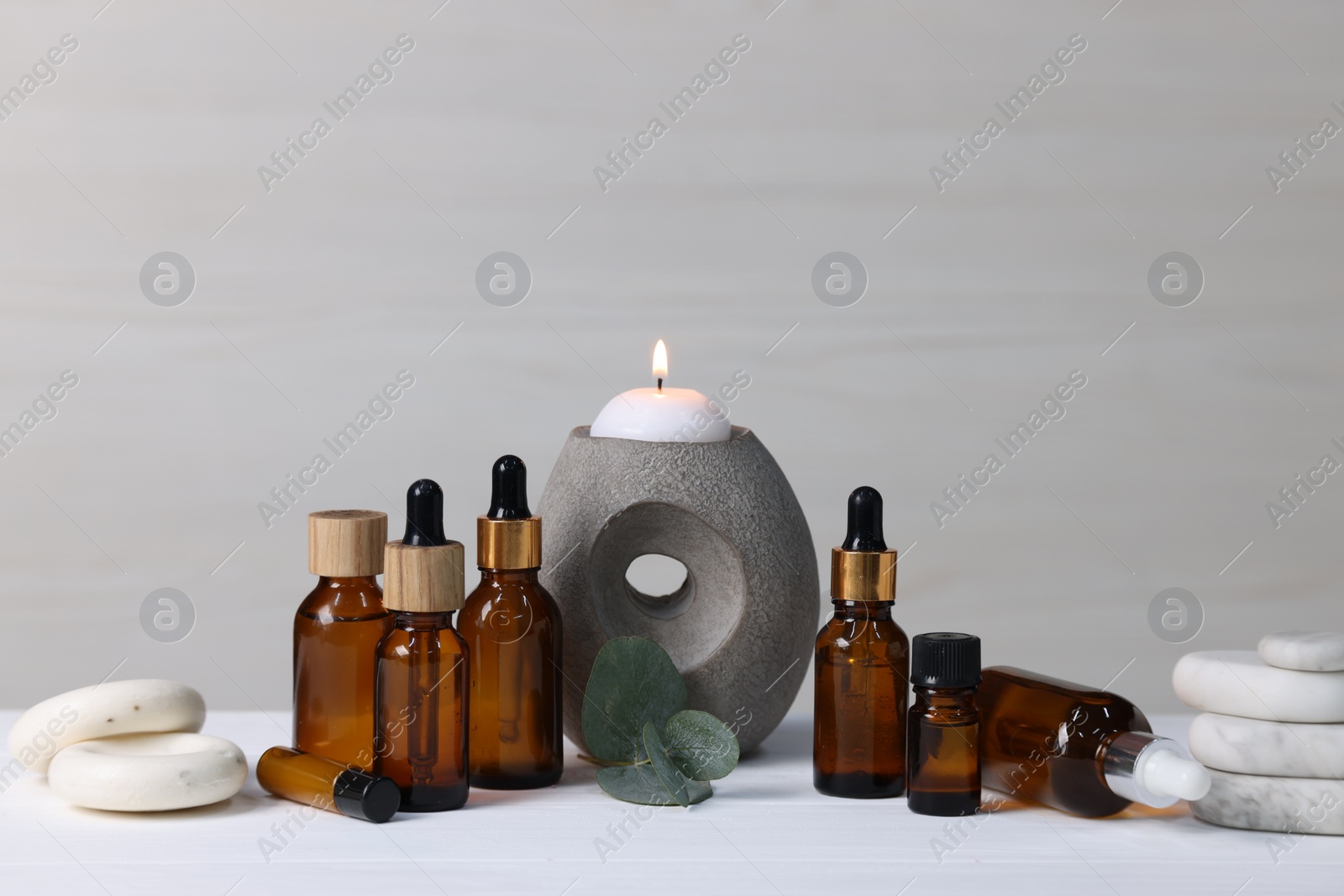 Photo of Different aromatherapy products, burning candle and eucalyptus leaves on white wooden table against light background