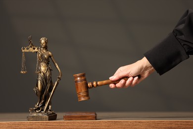 Judge striking mallet at wooden table against grey background, closeup. Figure of Lady Justice indoors