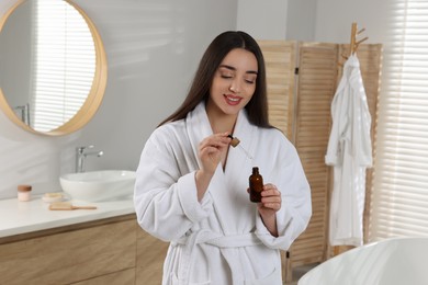 Photo of Happy young woman holding bottle and dropper of essential oil in bathroom