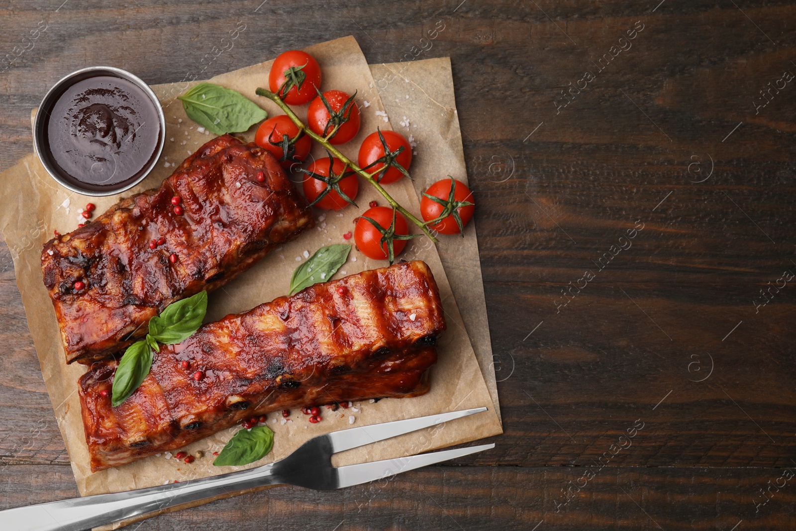 Photo of Tasty roasted pork ribs served with sauce, basil and tomatoes on wooden table, top view. Space for text
