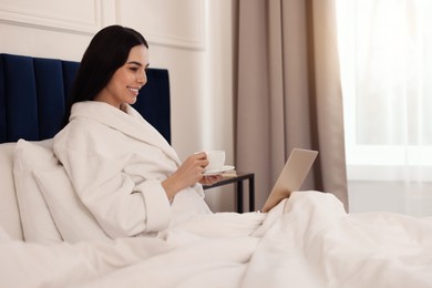 Photo of Happy young woman drinking coffee while working with laptop on bed in hotel room
