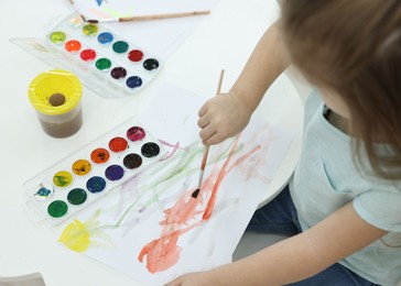 Photo of Little girl painting with brush and watercolor at white table, closeup