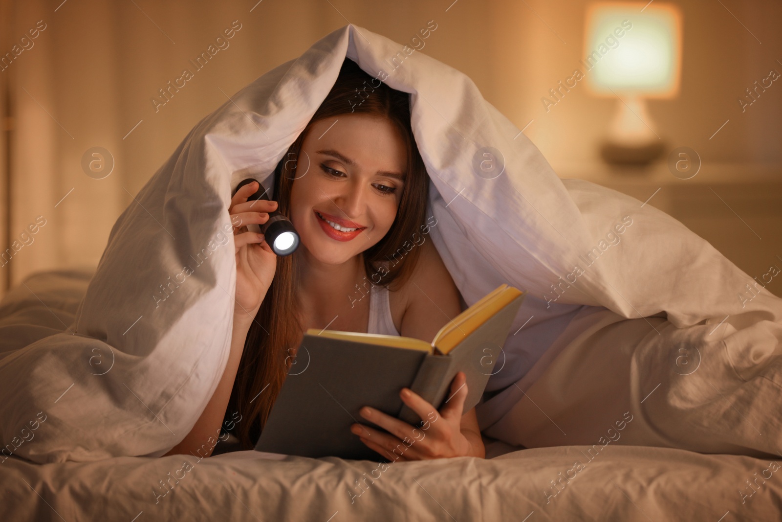 Photo of Young woman with flashlight reading book under blanket in bedroom