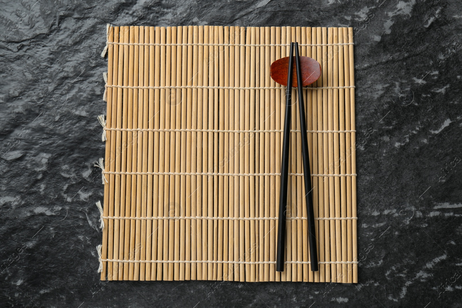 Photo of Bamboo mat with pair of chopsticks and rest on black textured table, top view