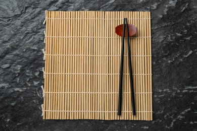 Photo of Bamboo mat with pair of chopsticks and rest on black textured table, top view