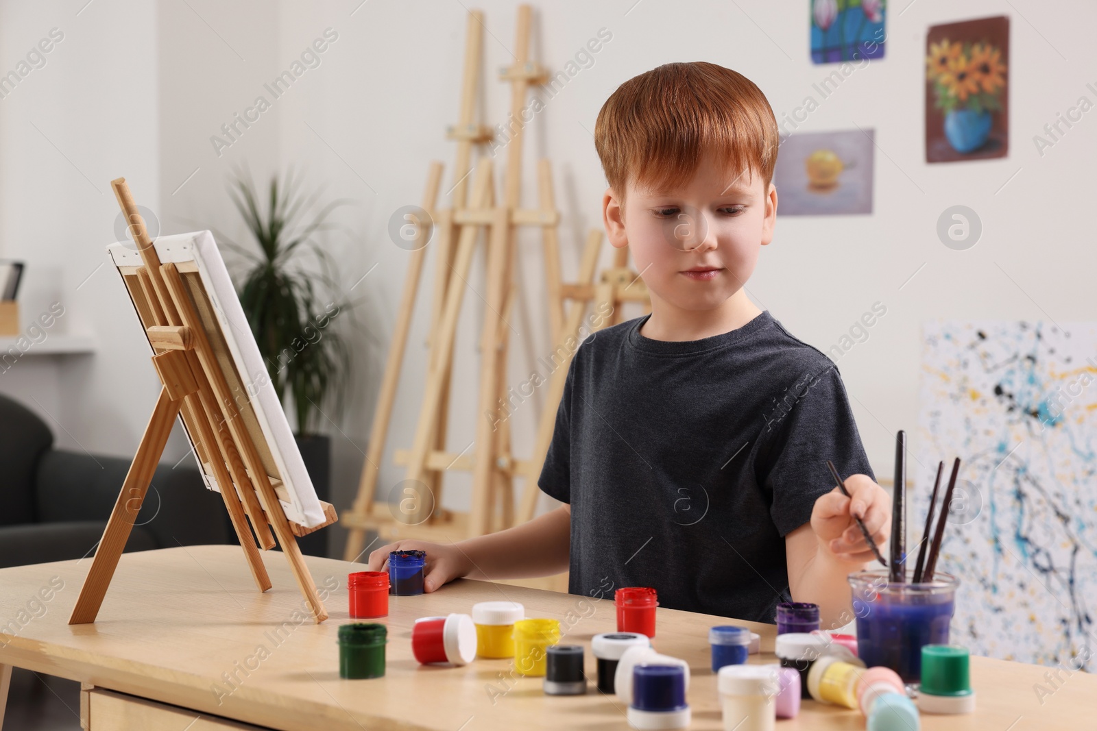 Photo of Little boy painting at table in studio. Using easel to hold canvas