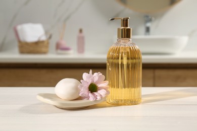 Photo of Glass dispenser with shower gel, soap bar and flower on white table in bathroom