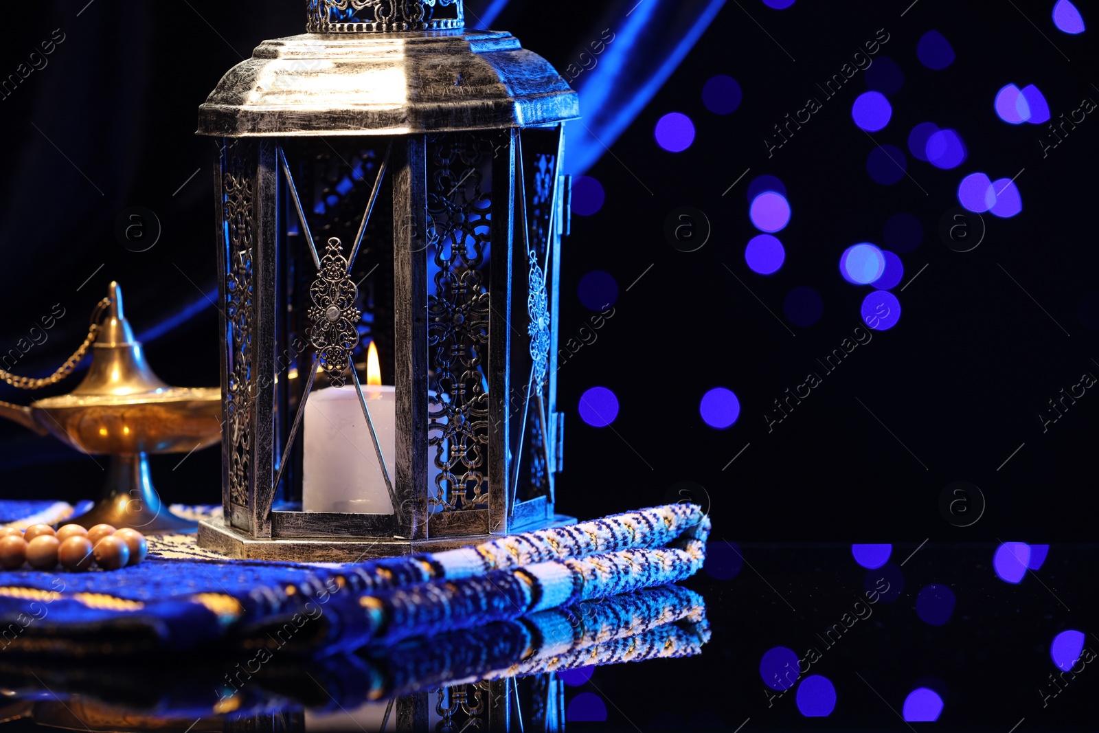 Photo of Arabic lantern, misbaha, Aladdin magic lamp and folded prayer mat on mirror surface against blurred lights at night. Space for text