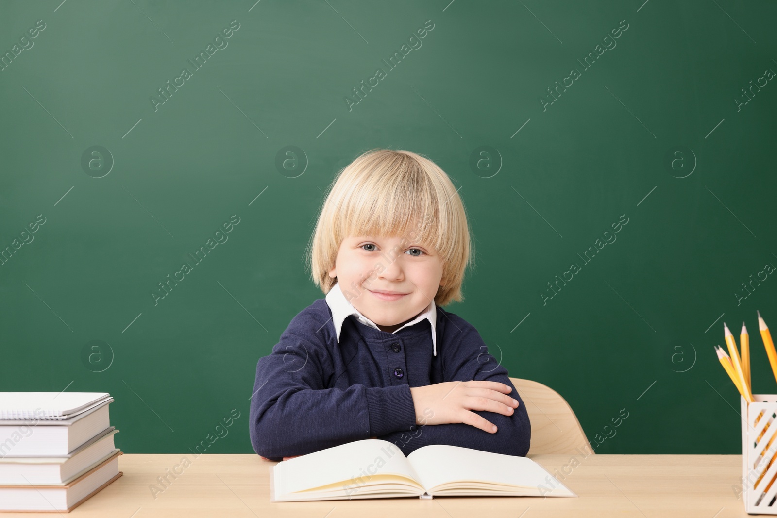 Photo of Happy little school child sitting at desk with books near chalkboard