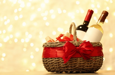 Photo of Festive basket with bottles of wine and gifts on table against blurred lights. Space for text