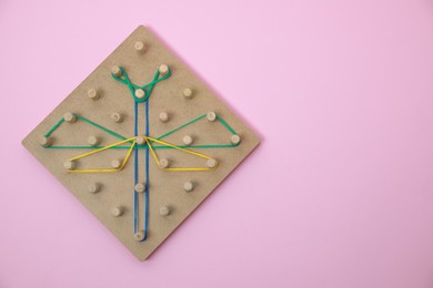 Photo of Wooden geoboard with dragonfly shape made of rubber bands on pink background, top view and space for text. Educational toy for motor skills development